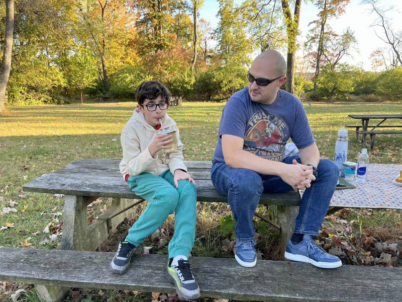 A kid and his dad sitting on a picnic table, the kid is drinking mate.